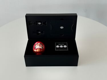 Eine ge&ouml;ffnete schwarze Geschenkbox zeigt eine rote Daruma-Puppe und ein Toyota-Logo auf einem Tisch.